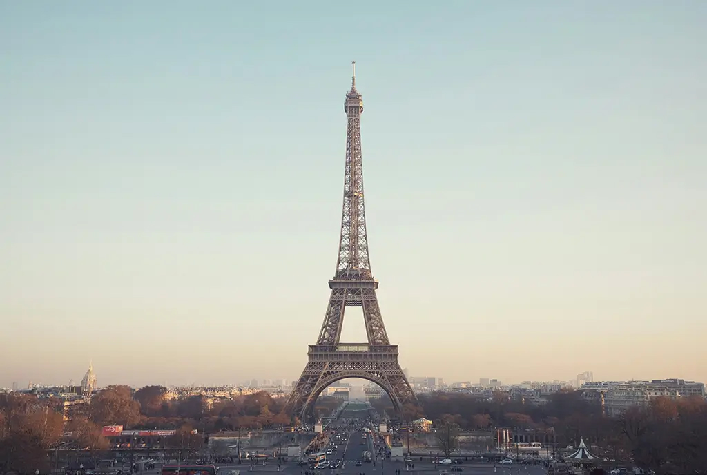 Der Eiffelturm in Paris, zentral vor einem leicht bew&ouml;lkten Himmel, umgeben von Geb&auml;uden und B&auml;umen im Vordergrund.